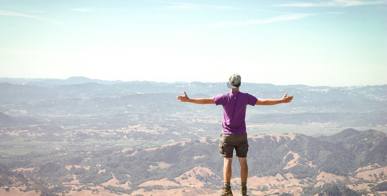 Man with wide open arms on top of a hill with wide open views in front him.